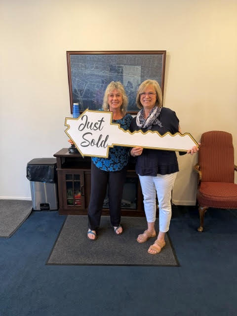 Two women holding a “Just Sold” sign in an office with office furniture and a corkboard backdrop