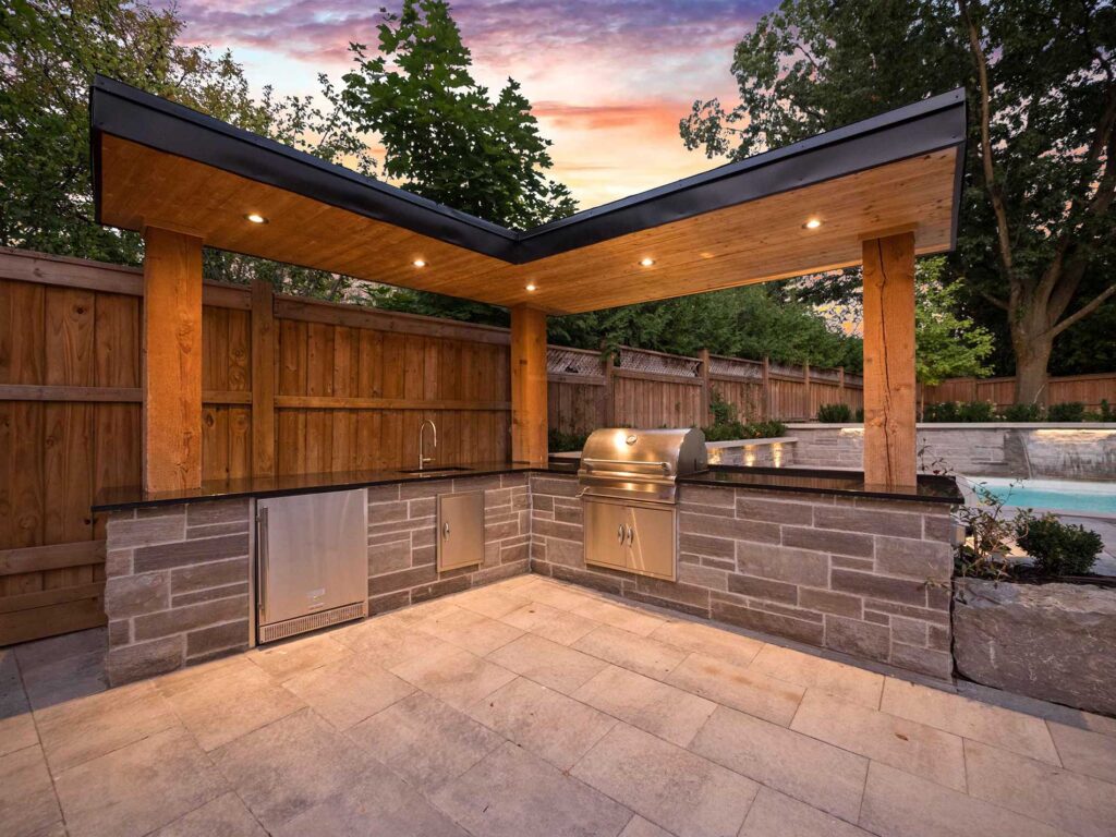 Outdoor kitchen with stone bar, luxury grill, black countertops and sink, large wood pillars hold roof overhang