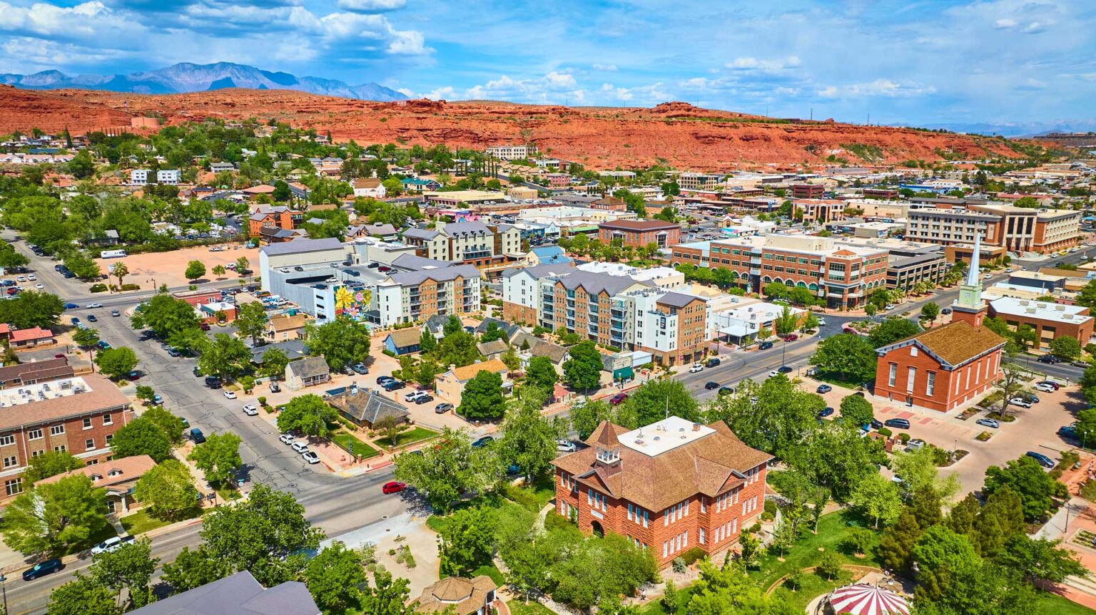 Aerial view of historic downtown St George Utah with red-brick landmarks lush green spaces and dramatic red rock mesas under clear blue skies showcasing vibrant city life and scenic desert landscape