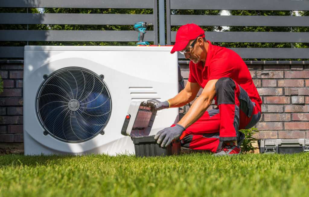 HVAC technician in red uniform servicing an outdoor heat pump unit on a grass lawn