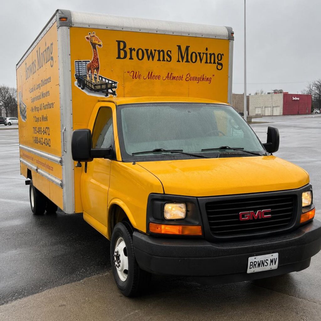Yellow Browns Moving GMC box truck parked on wet pavement on a rainy, overcast day. The truck features the Browns Moving logo, a giraffe mascot, and the slogan "We Move Almost Everything" on the side. Headlights are on, with commercial buildings visible in the background.
