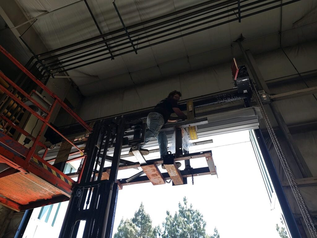 Pacific Overhead Door Service technician repairing an overhead garage door inside a shop with lift equipment